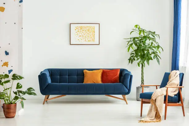 Photo of Mid-century modern chair with a blanket and a large sofa with colorful cushions in a spacious living room interior with green plants and white walls. Real photo.