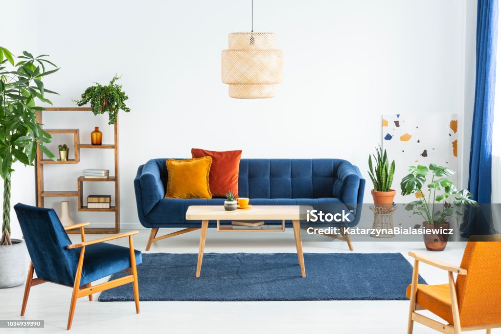 Retro armchairs with wooden frame and colorful pillows on a navy blue sofa in a vibrant living room interior with green plants. Real photo. Multi Colored Stock Photo