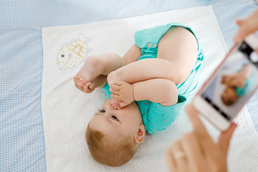 Parent taking photo of a baby with smartphone. Adorable newborn child taking foot in mouth. sucking feet. Digital family memories