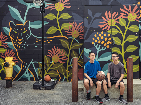 Twin brothers of Chinese  ethnicity posing with basketball while sitting at the alley basketball court featuring large mural on the wall behind. Urban city setting of North American city.