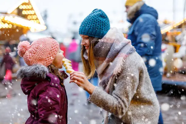 Photo of Young mother and daughter eating white chocolate covered fruits on skewer on traditional German Christmas market. Happy girl and woman on traditional family market in Germany, Munich during snowy day.