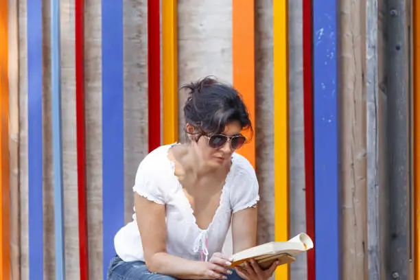 A woman in sunglasses sits on a wooden stage and reads a book