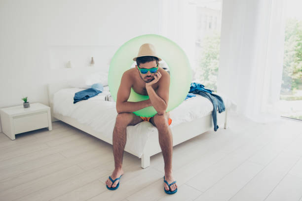 매력적인 잘생긴 젊은 수영 착용, 생활 부 표, 선글라스, 실내 침대에 앉아 슬픈 brunet 남자 수염. 바다 리조트 여름 휴가 - beach homes 뉴스 사진 이미지