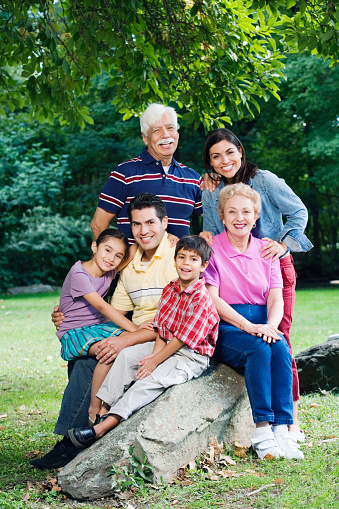 A happy family of four is spending time together outdoors. They are smiling and looking at the camera. The family is wearing casual clothes and is surrounded by trees and plants.