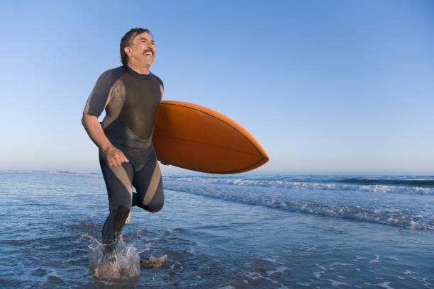 uomo ispanico che porta tavola da surf che corre lungo la spiaggia - senior adult surfing aging process sport foto e immagini stock