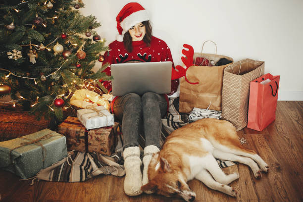 glückliches mädchen in weihnachtsmütze auf laptop arbeiten und mit niedlichen hund am goldenen schönen weihnachtsbaum mit lichtern und geschenke in festlicher raum sitzen. stimmungsvolle momente mit der familie. freelance - christmas shopping internet family stock-fotos und bilder