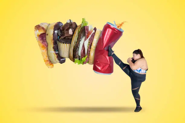 Picture of strong fat woman kicking soft drink and fast food. Shot with yellow background