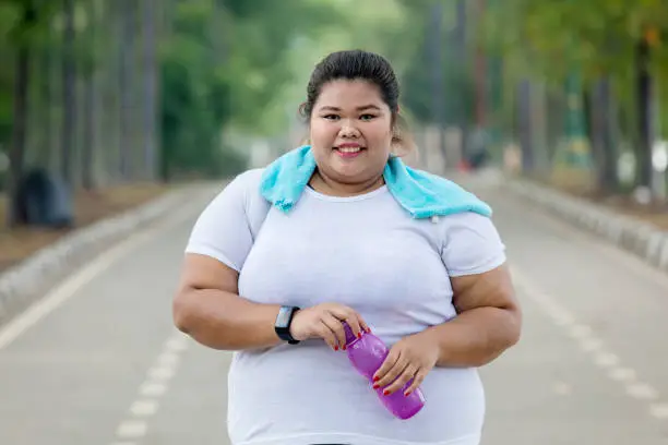 Portrait of fat woman holding a bottle of water and resting after jogging on the road