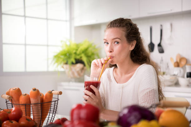 mulher jovem e saudável em uma cozinha com frutas e legumes e suco - dieting juice carrot tomato - fotografias e filmes do acervo