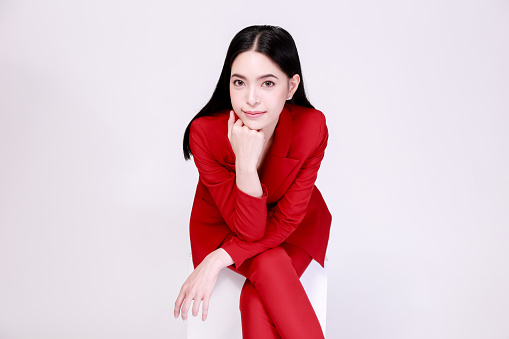 Young confident Asian woman in red elegant suit with pants posing sitting in half body isolated over white background