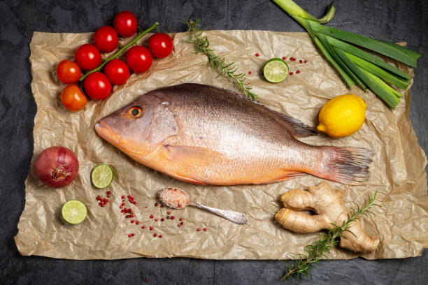 Fresh raw red snapper fish with spices , herbs and vegetables for cooking on brown backing paper . Top view. stock photo