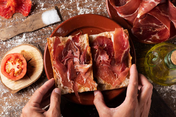 spanish ham sandwich, serrano ham sandwich high angle view of a man preparing a typical spanish bocadillo de jamon, a serrano ham sandwich, on a rustic wooden table, next to a plate with some slices of serrano ham and a cruet with olive oil cold cuts meat photos stock pictures, royalty-free photos & images