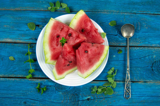pieces of ripe watermelon on a white plate, decorated with fresh mint and a spoon on a blue background stock photo