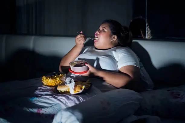 Unhealthy lifestyle concept: Overweight woman eating junk food in bed before sleeping
