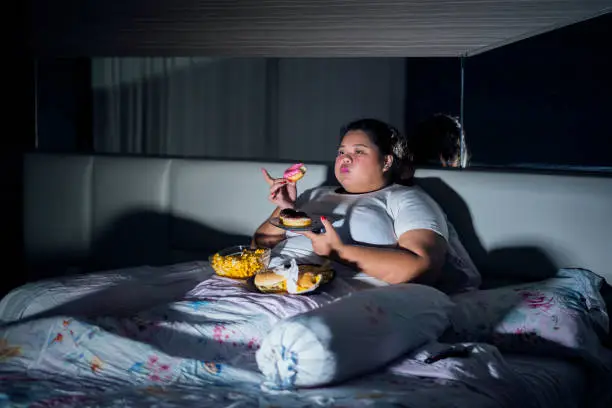 Unhealthy lifestyle concept: Overweight woman eating ice cream and junk food in bed before sleeping