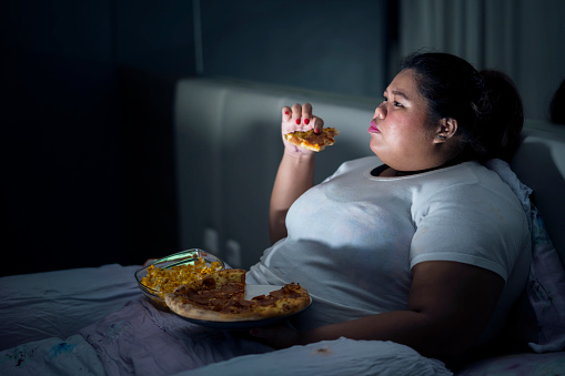 Fat woman eating pizza on bed while watching movie before sleep