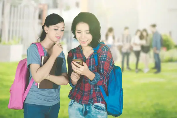 Picture of two female college students using a smartphone together while standing in the park