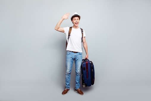 Hey, I returned from vacation! Full-size portrait of guy with a suitcase and a backpack, who waving his hand and look at the camera isolated on light gray background