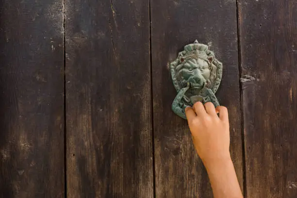 Photography of a child's hand, knocking on an old wooden door with a metal door knocker, ideal for memes, notes, posters, background, texture, wallpaper, etc. The photography is a close up of the pattern from near distance. It's shot in daylight. The rusty door handle is antique and the wood is old and structured. There are four stripes of dark brown wood. The wooden gate is well preserved. The metal door knock handle is in the right corner of the photography, it has a human face and is a beautiful ornament for decoration.. The shot is taken in the Old Town of Nessebar, Burgas, Bulgaria. The girl's arm is stretched from the bottom of the shot.