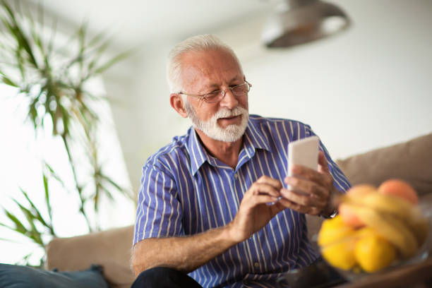 anciano con un smartphone y sonriendo mientras está sentado en el sofá en casa - surfista de plata fotografías e imágenes de stock