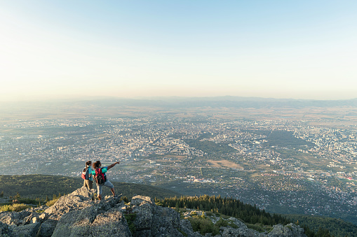 Young couple hikers standing on the mountain peak, looking and pointing at the city in the valley below