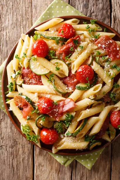 Photo of Delicious hot penne pasta with ham, cherry tomatoes, zucchini and cheese close-up. Vertical top view