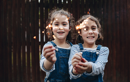 Two little girls holding sparkling hand fireworks. Identical twin sisters burning sparklers outdoors and smiling.
