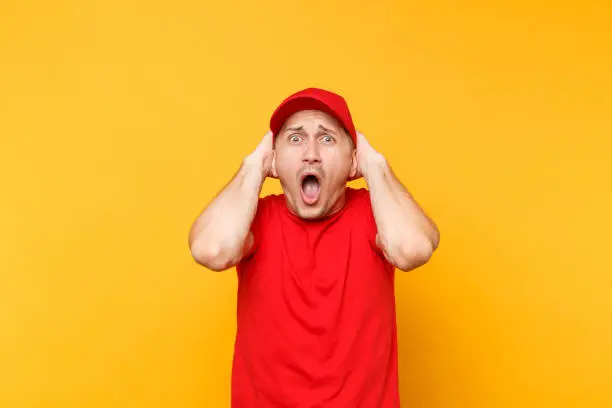 Photo of Delivery man in red uniform isolated on yellow orange background. Professional perturbed shocked male employee in cap, t-shirt working as courier dealer put hands on head. Service concept. Copy space.