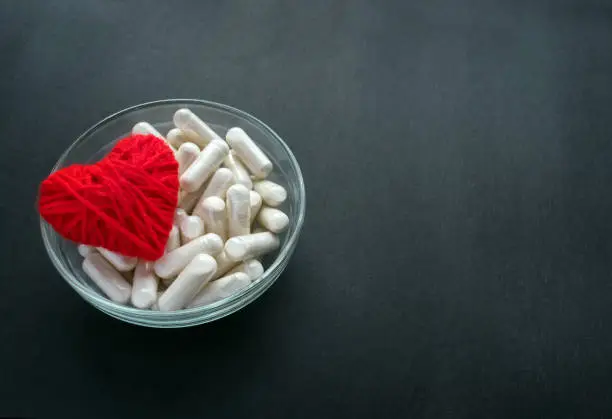 Photo of White veg capsules and red thread heart in glass bowl on black background. Anticoagulant, Blood Thinners. Cardiac Medications, pills for the heart. Treat Heart disease Failure