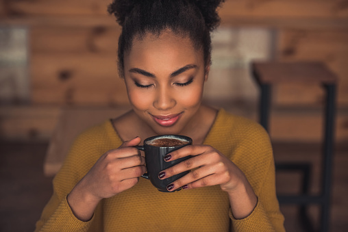 Beautiful Afro American girl in casual clothes is smelling coffee and smiling while resting in cafe