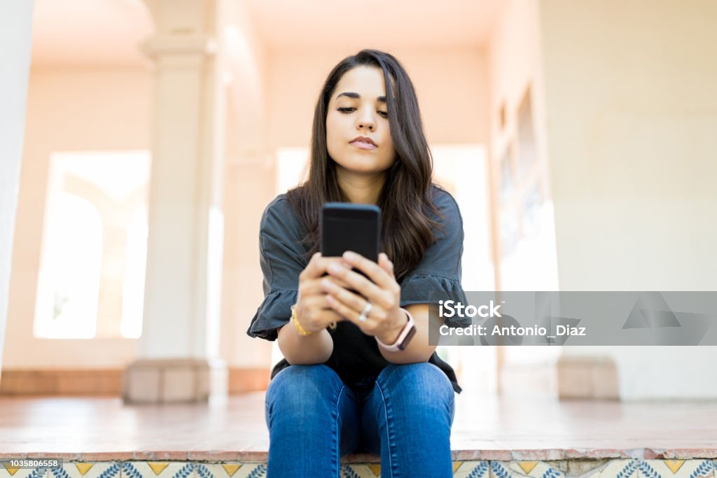 Woman Checking Email On Modern Cellular While Sitting At Corridor Mid adult woman in casuals checking email on modern cellular while sitting at corridor 30-39 Years Stock Photo