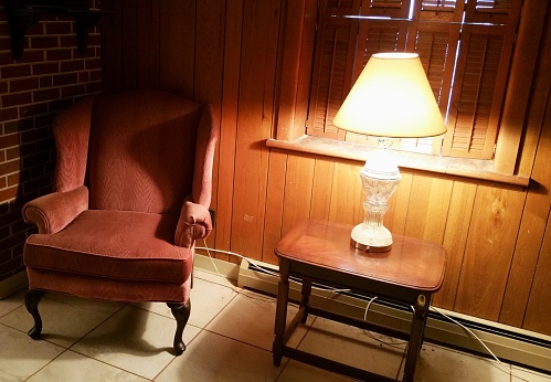 Mauve-colored, high back armchair, located in corner of home office, with nearby lamp on top of small table.  Wood paneling.  Window with closed, wooden shutters.  White tile floor.