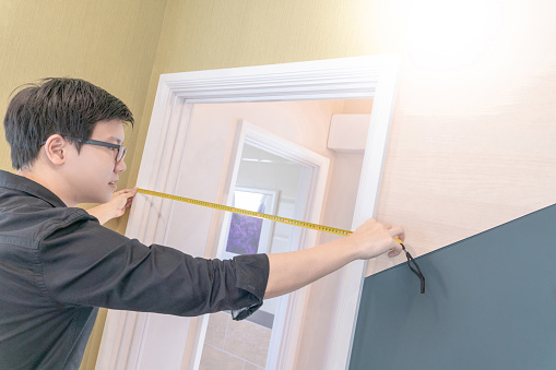 Young Asian worker man using tape measure on door frame for door installation. Home interior designer. Housing design and construction concept