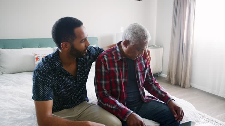 Adult son comforting depressed senior father sitting in bedroom at home - shot in slow motion