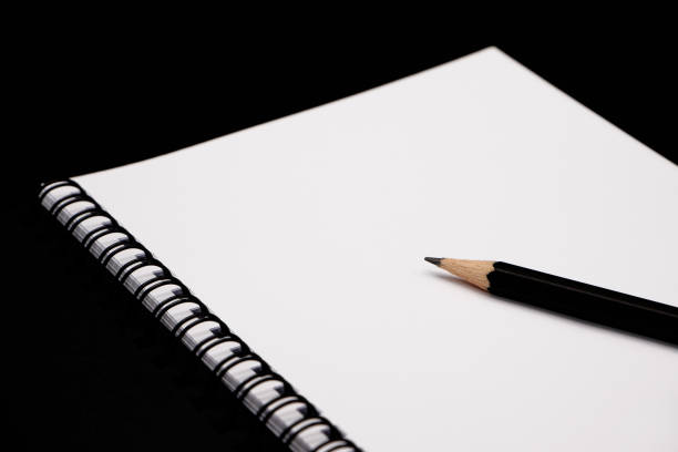Black pencil on opened black spiral notebook with blank white page on black background.