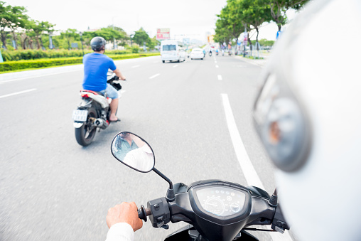 Da Nang City, Vietnam - May 7, 2018: View from behind the motorcycle driver's back. White helmet, a hand, motorbike's dashboard and the street with another scooter (blurred) going ahead towards other traffic. Traveling by motor bike taxi.