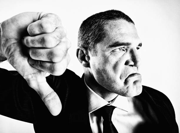 A furious businessman, shot in black and white and distorted by fisheye lens, frowns and gives an emphatic, disapproving, thumbs-down signal.
