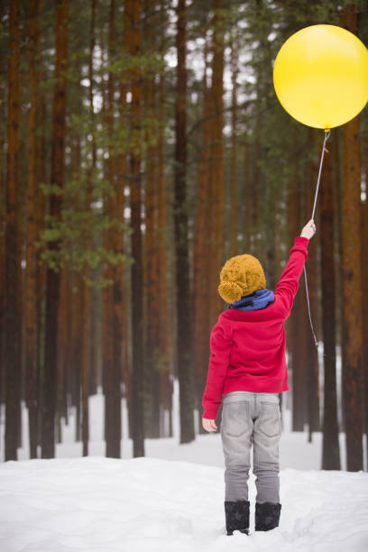 маленький счастливый мальчик в джинсах и красном свитере стоит в зимнем лесу и держит в руках большой желтый воздушный шар. день рождения м� - balloon child winter snow стоковые фото и изображения