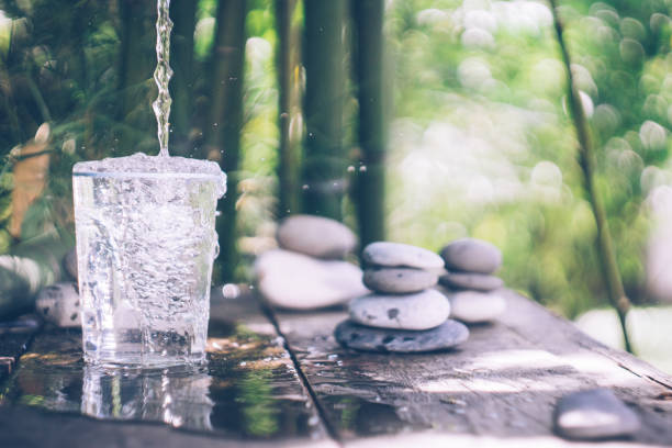 чистая вода льется в стекло рядом с камнями на старом деревянном столе среди бамбука - spa treatment health spa zen like bamboo стоковые фото и изображения
