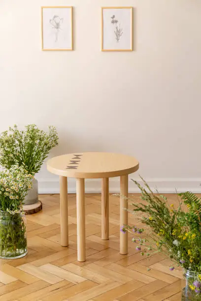 A small round wooden table surrounded by fresh meadow flowers standing on a parquet against a light beige wall with illustrations of nature in a sunny guestroom interior. Real photo.
