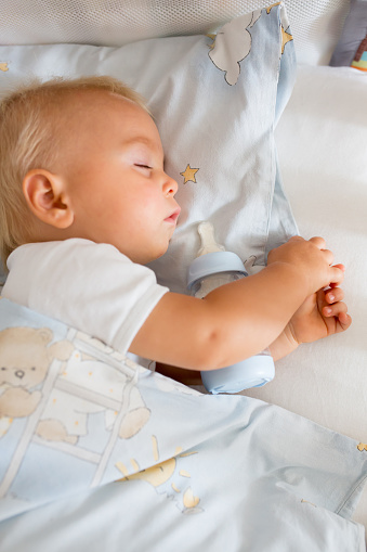 Cute little baby boy, sleeping with bottle with formula milk. Tired child in baby cot bed