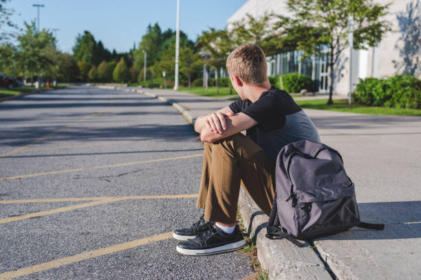 Lonely teenage boy sitting on curb next to high school. The image displays a lonely teenage boy sitting on the curb in front of his high school at the end of the school day. homelessness stock pictures, royalty-free photos & images
