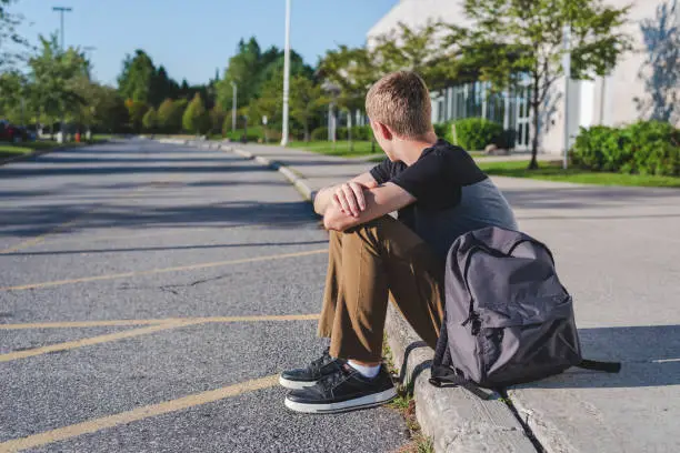 Photo of Lonely teenage boy sitting on curb next to high school.