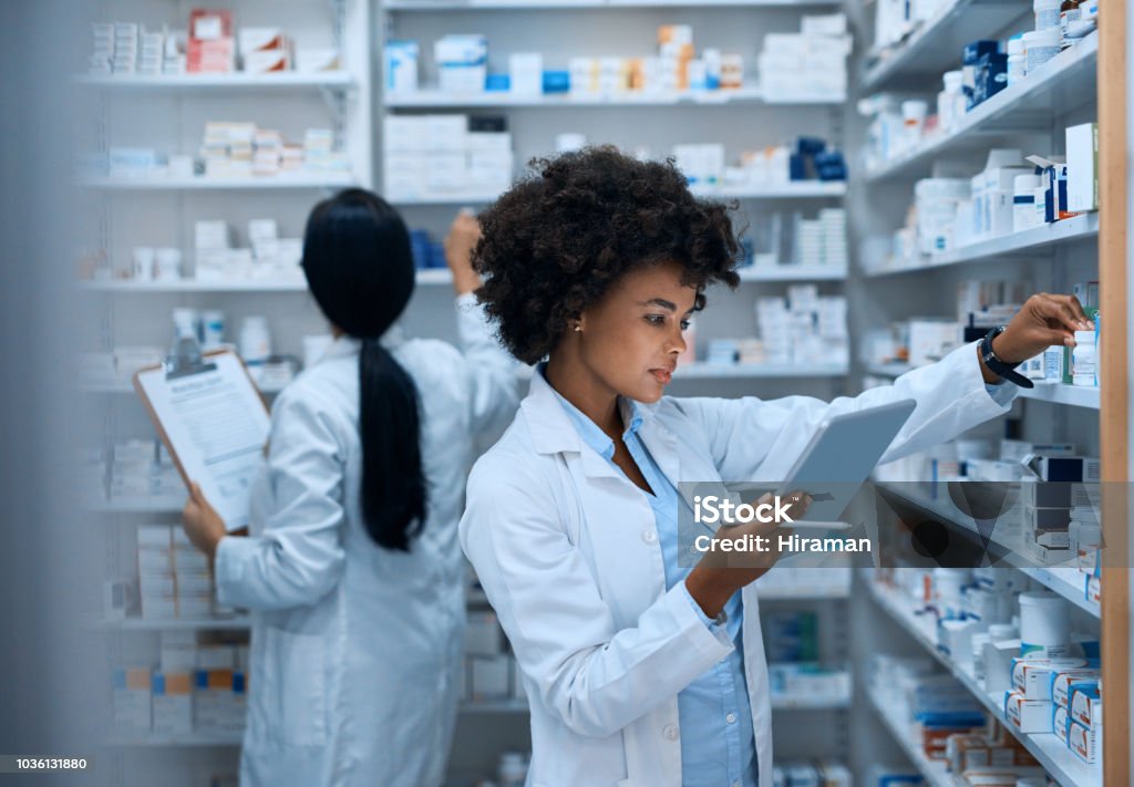 Efficient pharmacy operations thanks to teamwork Shot of a young woman doing inventory in a pharmacy on a digital tablet with her colleague in the background Pharmacy Stock Photo