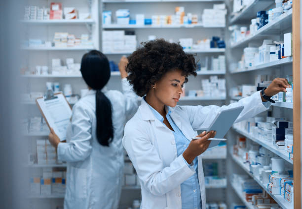 Efficient pharmacy operations thanks to teamwork Shot of a young woman doing inventory in a pharmacy on a digital tablet with her colleague in the background african american scientist stock pictures, royalty-free photos & images