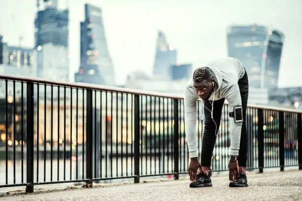 Photo of Black man runner with smartphone in an armband on the bridge in a city, stretching.