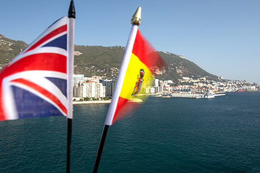 The Union Jack and the Spanish Flag fluttering in the wind with Gibraltar in the background, indicating the dispute over its sovereignty and the effects of Brexit