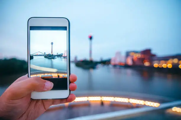 A human hand holding a mobile phone in order to take a picture of Düsseldorf, Germany, at night