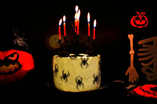 Photo of HAPPY HALLOWEEN CAKE PARTY. DECORATED WITH SPIDERS AND RED BURNING CANDLES AND A CHANDELIER AGAINST BLACK COLORED BACKGROUND AND SOME PUMPKINS ANS A SKULL.