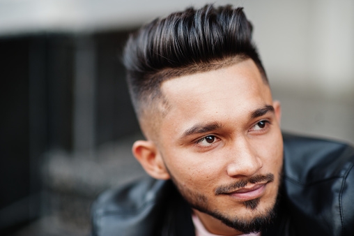 Stylish indian beard man at black leather jacket. India model posed sitting at streets of city. Close up portrait.